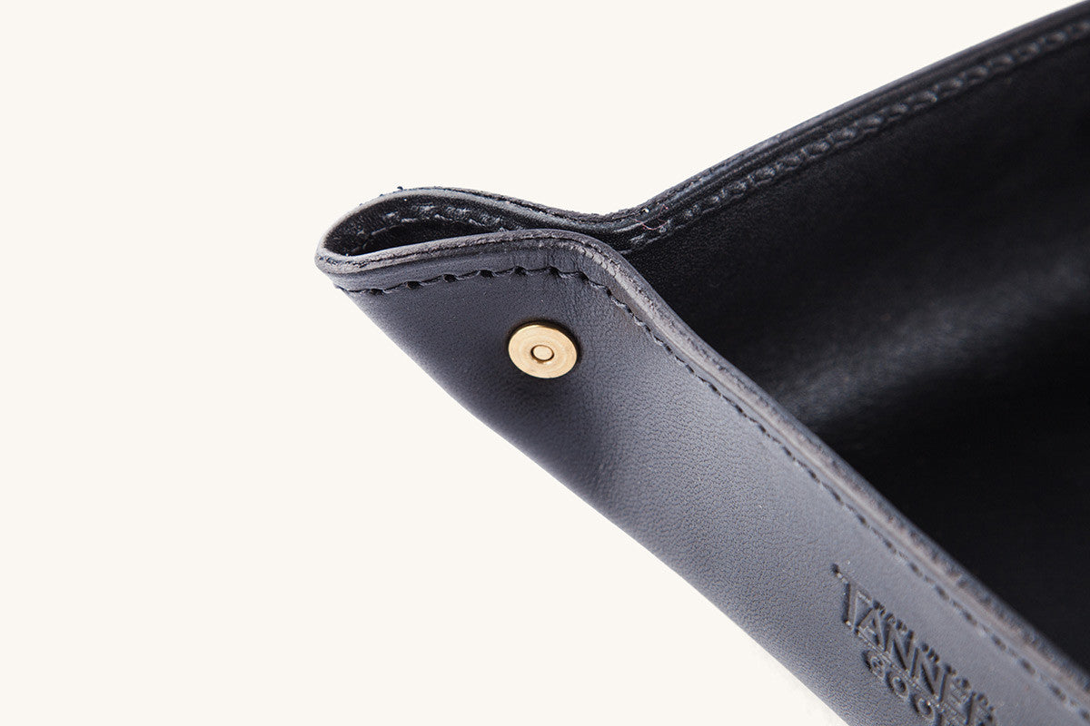 Angled view of a black leather valet tray with a brass corner snap, visible stitching, and debossed text.