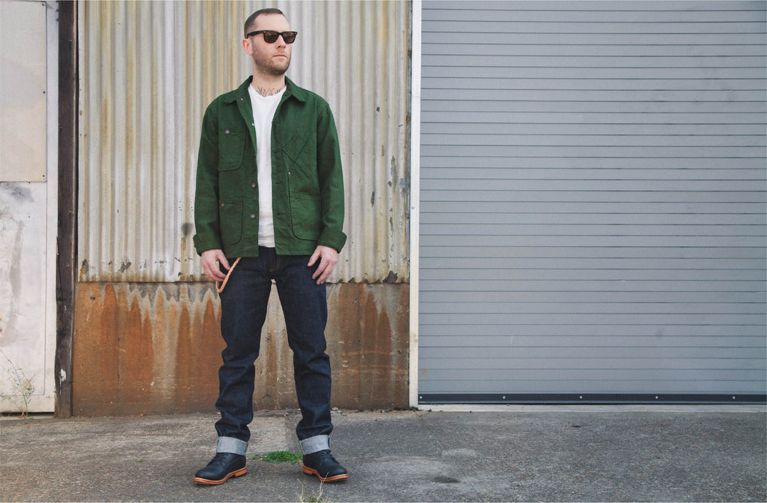 Man in green jacket and dark jeans stands confidently against rusted metal wall, exuding rebel style.
