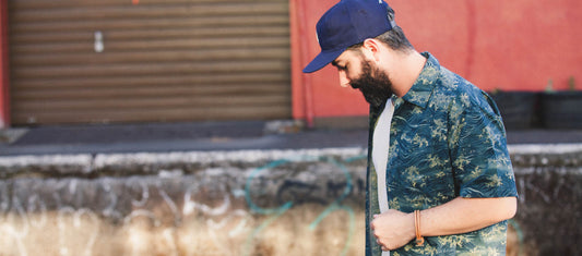 Bearded man in blue cap and patterned shirt walks thoughtfully past graffiti-covered wall, embodying urban calm.