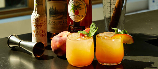 Two vibrant peach cocktails garnished with mint and peach slices, surrounded by bottles of bourbon, peach liqueur, and bitters on a sunlit bar.