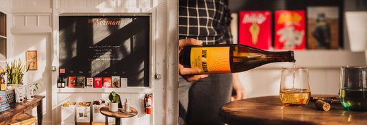 Person pouring golden wine into glass at Bar Norman, cozy shop with chalkboard sign and colorful art.