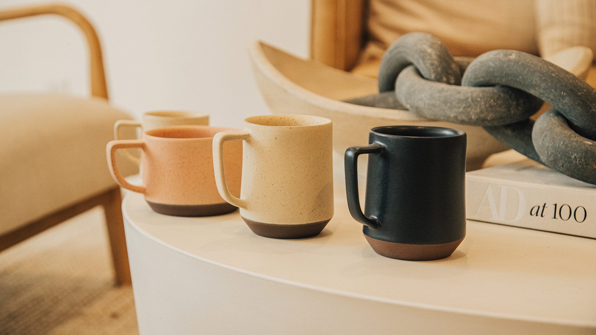 Earthy ceramic mugs in terracotta, cream, and black on a minimalist table, styled with a sculptural chain and design book.
