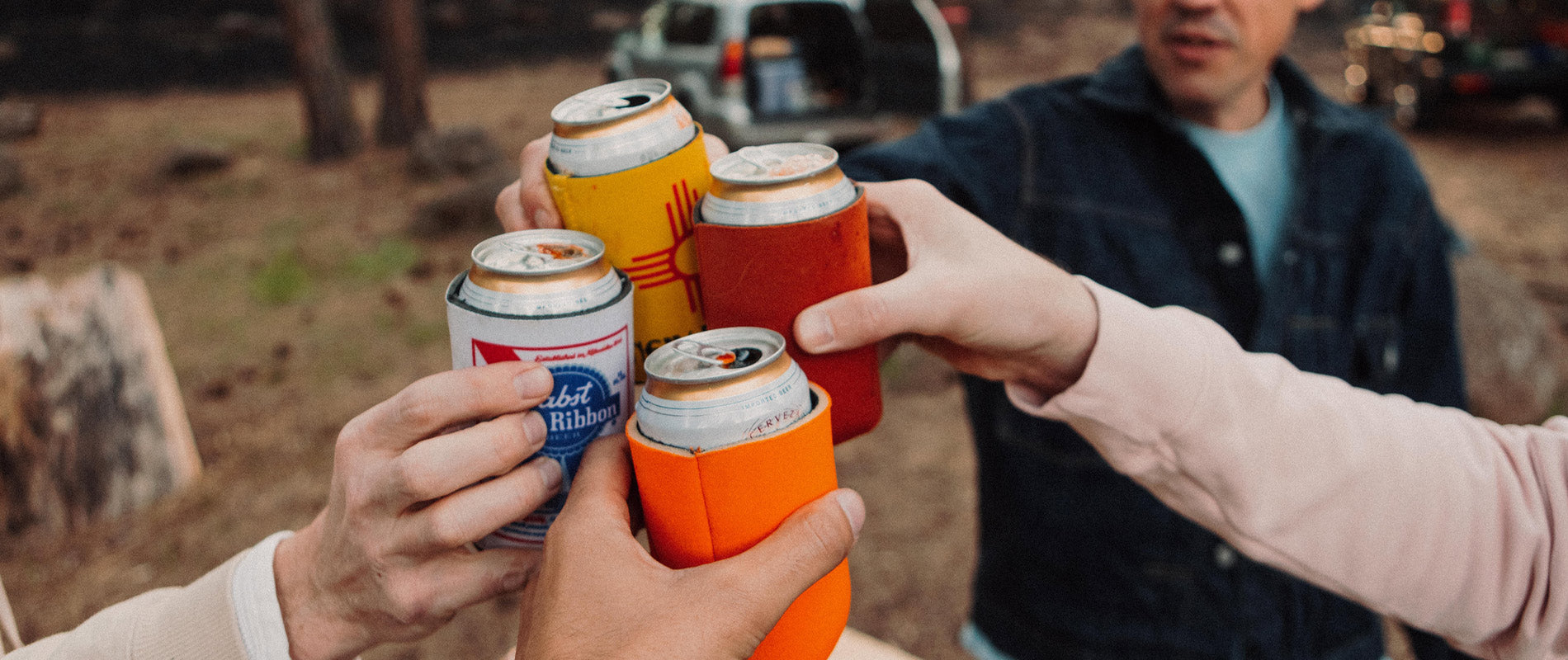 Hands toasting with colorful koozies in forest setting, celebrating Memorial Day weekend.