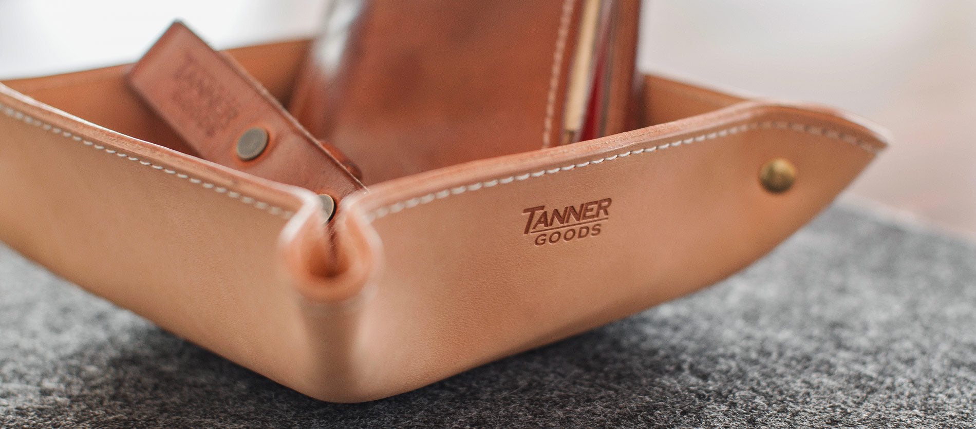 Rich tan leather valet tray with white stitching, brass hardware, and Tanner Goods branding, holding pens and cards on textured gray surface.