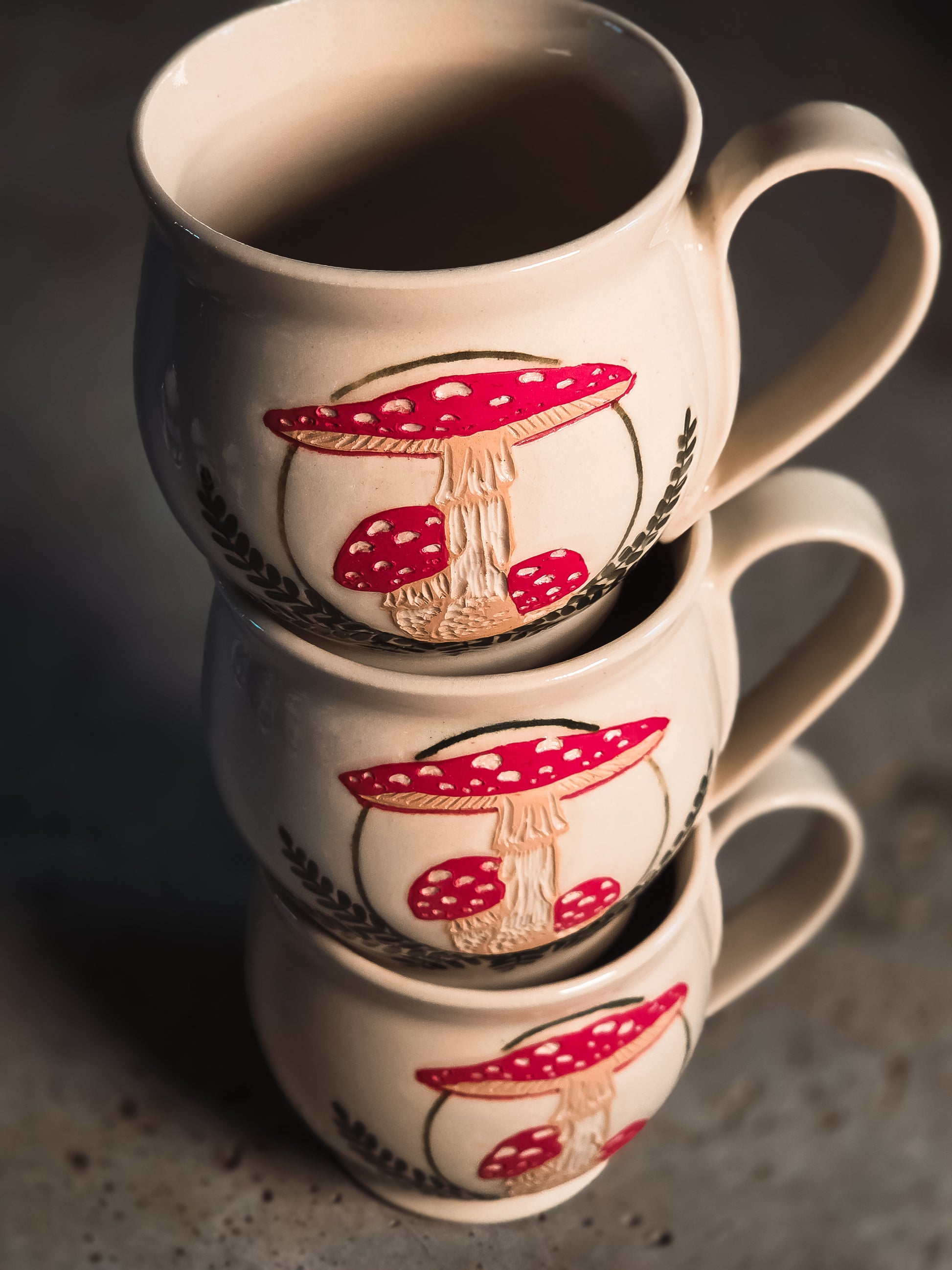 Cream ceramic mugs stacked, decorated with vibrant red and white Amanita mushroom designs.
