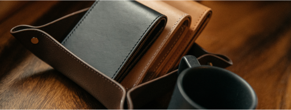 Brown leather wallet open on wooden surface, holding black cardholder, beside dark mug, warm lighting.
