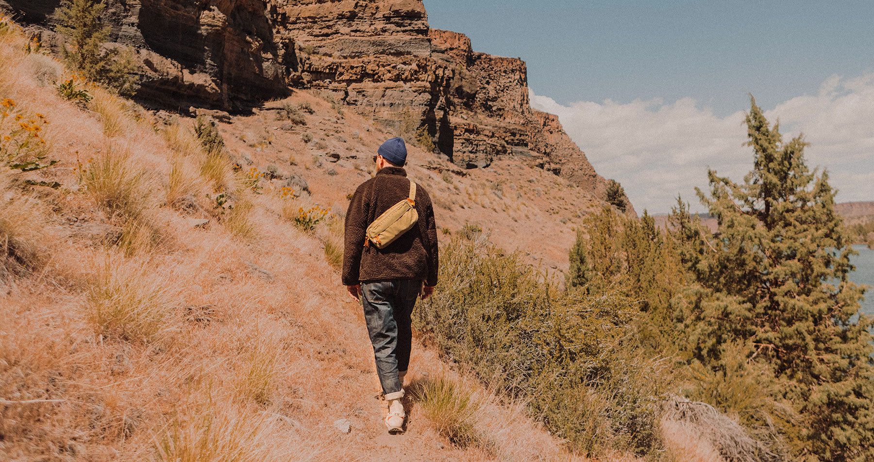 Person in blue beanie and brown jacket hikes along dry trail, red rock cliffs and green trees in background under partly cloudy sky.