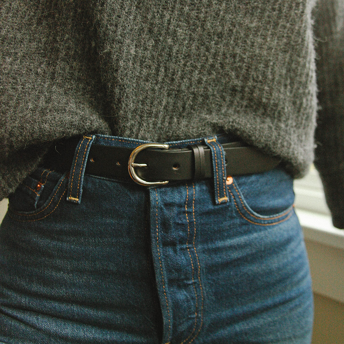 Black leather belt with a silver D-shaped buckle, styled with blue jeans and a chunky grey sweater.