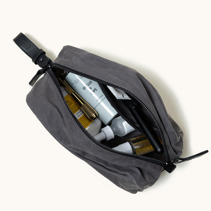 Slate grey waxed canvas Drifter Dopp Kit, open with various toiletries, black zipper, and leather strap.